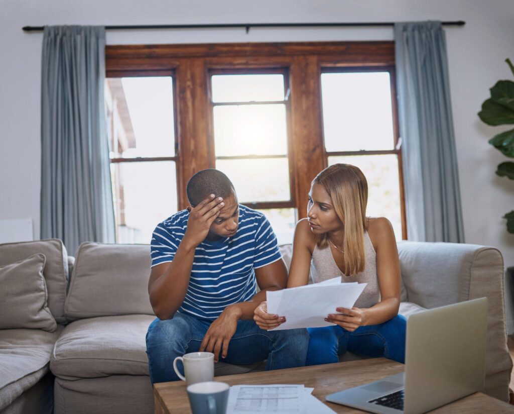 Couple reviewing bills and paperwork with concerned expressions while sitting on a sofa.